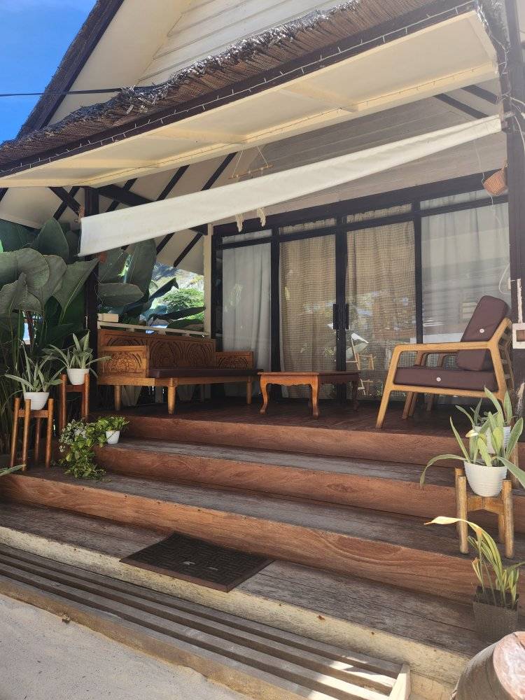 A charming entrance features a wooden staircase leading to a covered porch. Seating arrangements include a wooden table and an armchair, surrounded by potted plants. Large glass doors provide a view into the interior, with a canopy overhead for shading.