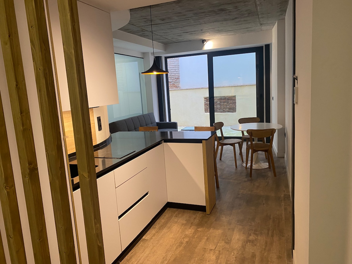 A modern kitchen area features sleek cabinetry and a spacious countertop. A dining table with wooden chairs is positioned nearby, complemented by a window that allows natural light to fill the space. The living area, visible through glass doors, includes a comfortable sofa.