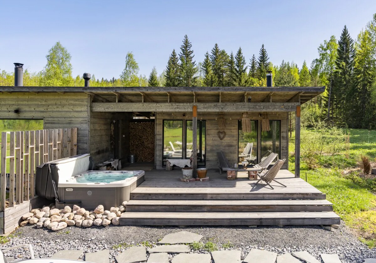A wooden exterior reveals a spacious deck featuring lounge chairs and a hot tub. Large glass doors provide access to the interior, while natural scenery of trees and a grassy landscape creates a peaceful environment.
