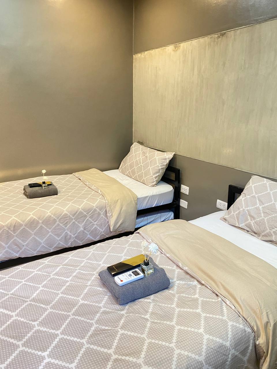 The room displays two single beds, each topped with neutral-toned bedding and decorative pillows. A small nightstand is positioned between the beds, featuring a candle and guest kit. The walls are painted in a soft gray, enhancing the room's calming atmosphere.