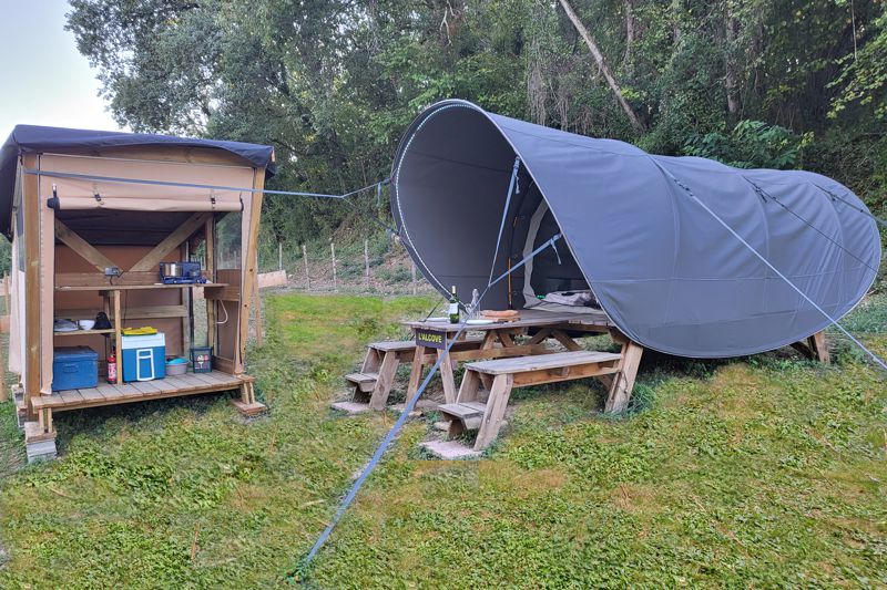 An outdoor camping setup includes a spacious tent and a covered kitchenette on a wooden platform. The kitchenette features a gas stove and storage for dishware. A picnic table with benches is positioned nearby, surrounded by green grass and trees.