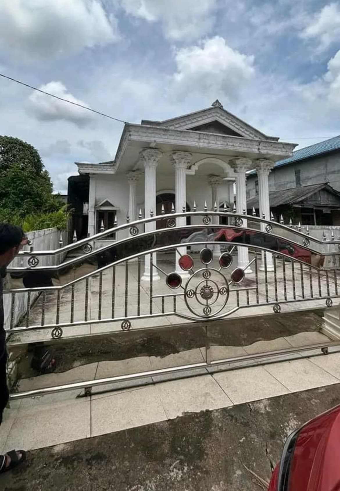 An ornate entrance gate features intricate metalwork and decorative elements. Beyond the gate, a unique house showcases classical architecture with tall columns supporting a pediment. A spacious courtyard is visible, bordered by paving stones and lush greenery under a partly cloudy sky.