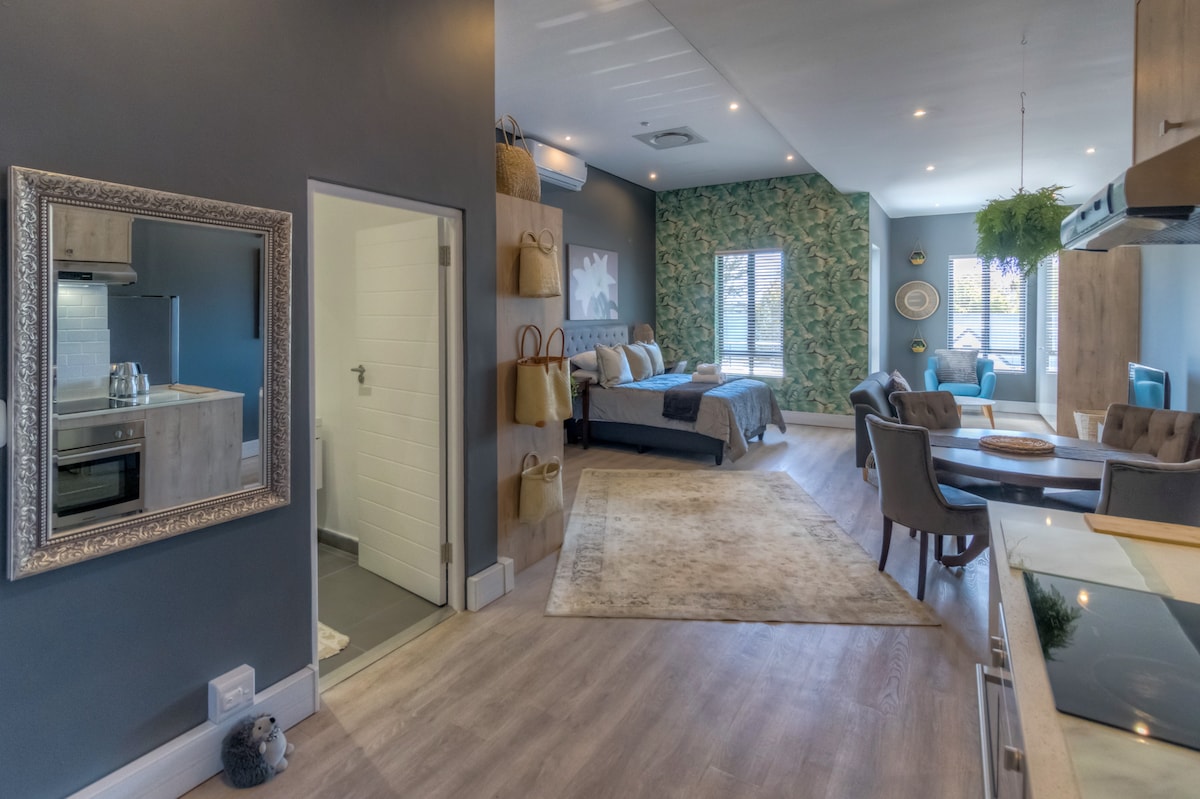An inviting studio apartment interior is displayed, featuring a modern kitchen area with upscale appliances. A cozy sleeping area with patterned wallpaper is visible, alongside a dining space. Natural light fills the room through large windows, complemented by a decorative mirror and neutral-toned rug.