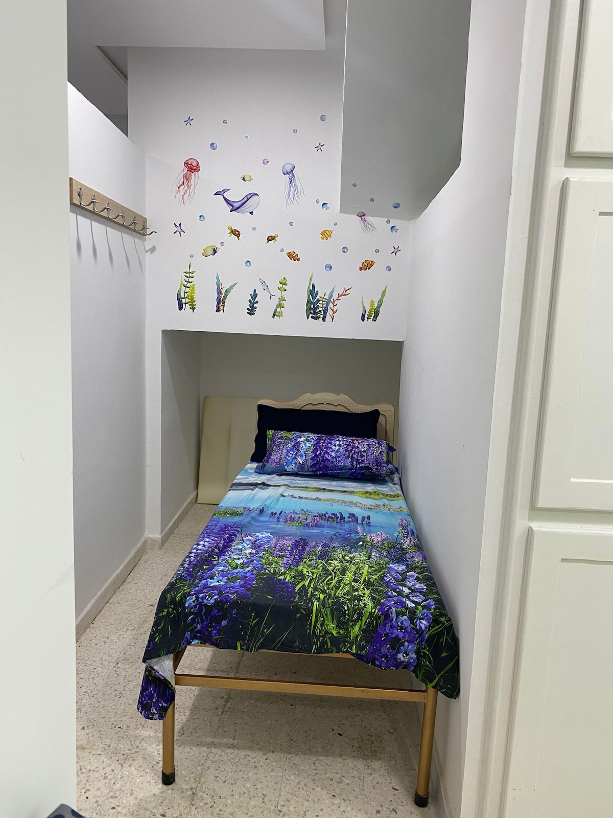A narrow sleeping space features a single bed adorned with a floral bedspread in shades of blue and purple. The wall above displays a whimsical mural of underwater life, while a row of hooks is positioned nearby for convenience.