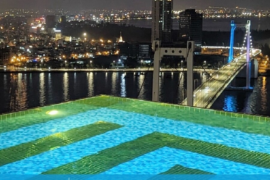 An expansive rooftop pool reflects city lights at night, with vibrant turquoise water creating a welcoming ambiance. In the distance, a lit bridge connects both sides of the waterfront, enhancing the skyline view.