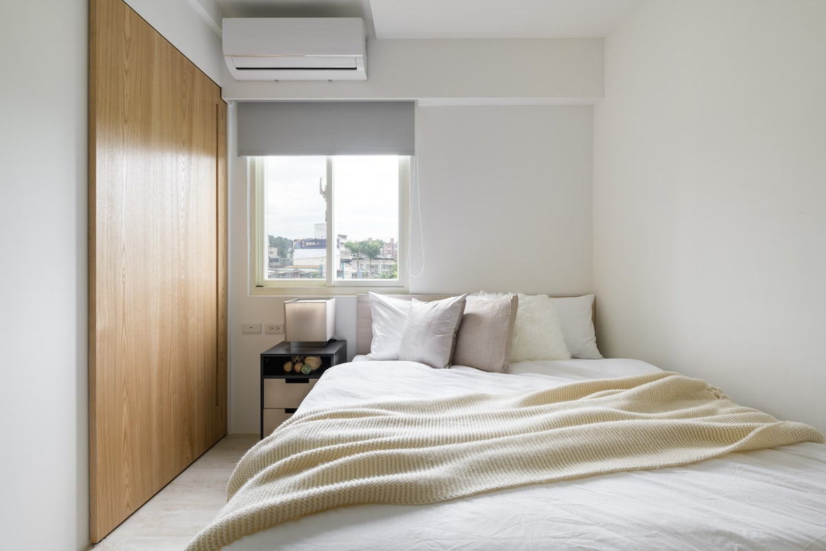 A bright bedroom features a spacious bed topped with soft pillows and a light blanket. Natural light filters through a window dressed with a simple shade. A nightstand with decorative items is positioned beside the bed, and a closed wooden door adds privacy.