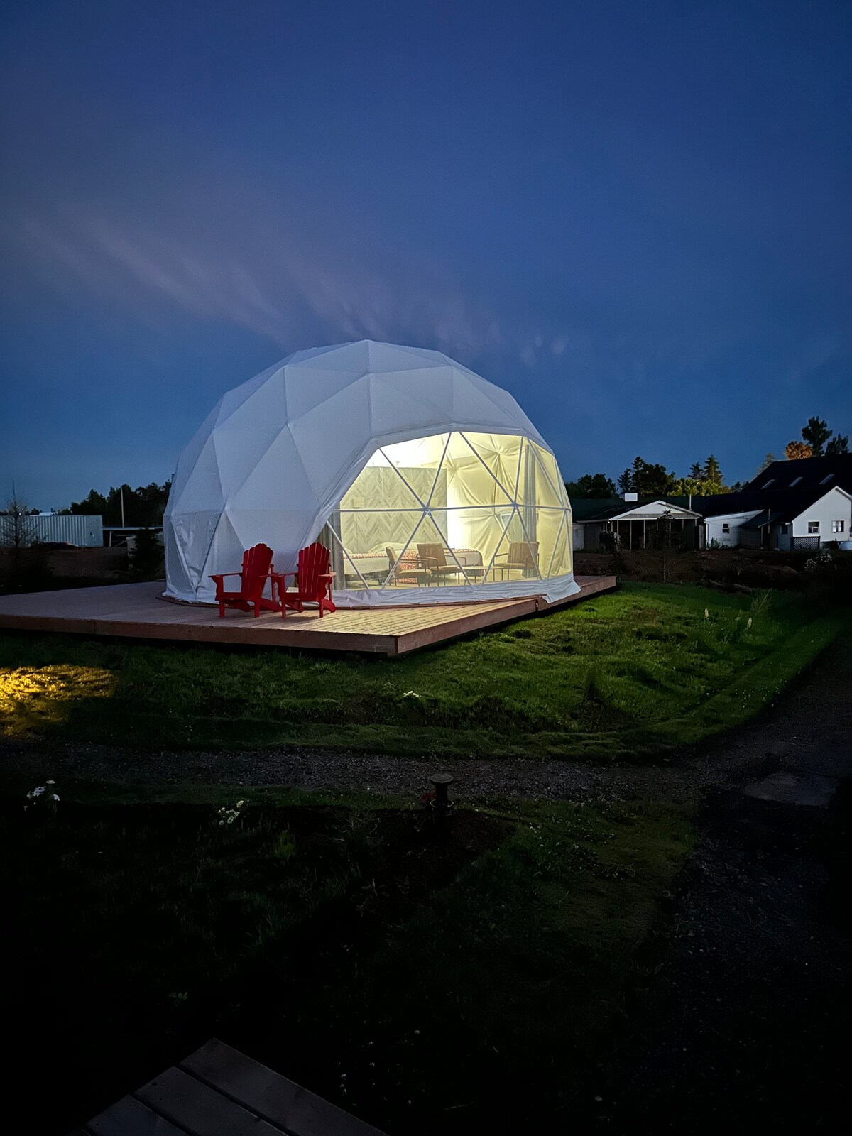A geodesic dome is illuminated from within, showcasing its unique structure against the evening sky. Red lounge chairs are positioned on the wooden deck, inviting relaxation. Soft lighting highlights the surrounding grass and pathway, enhancing the sense of tranquility.