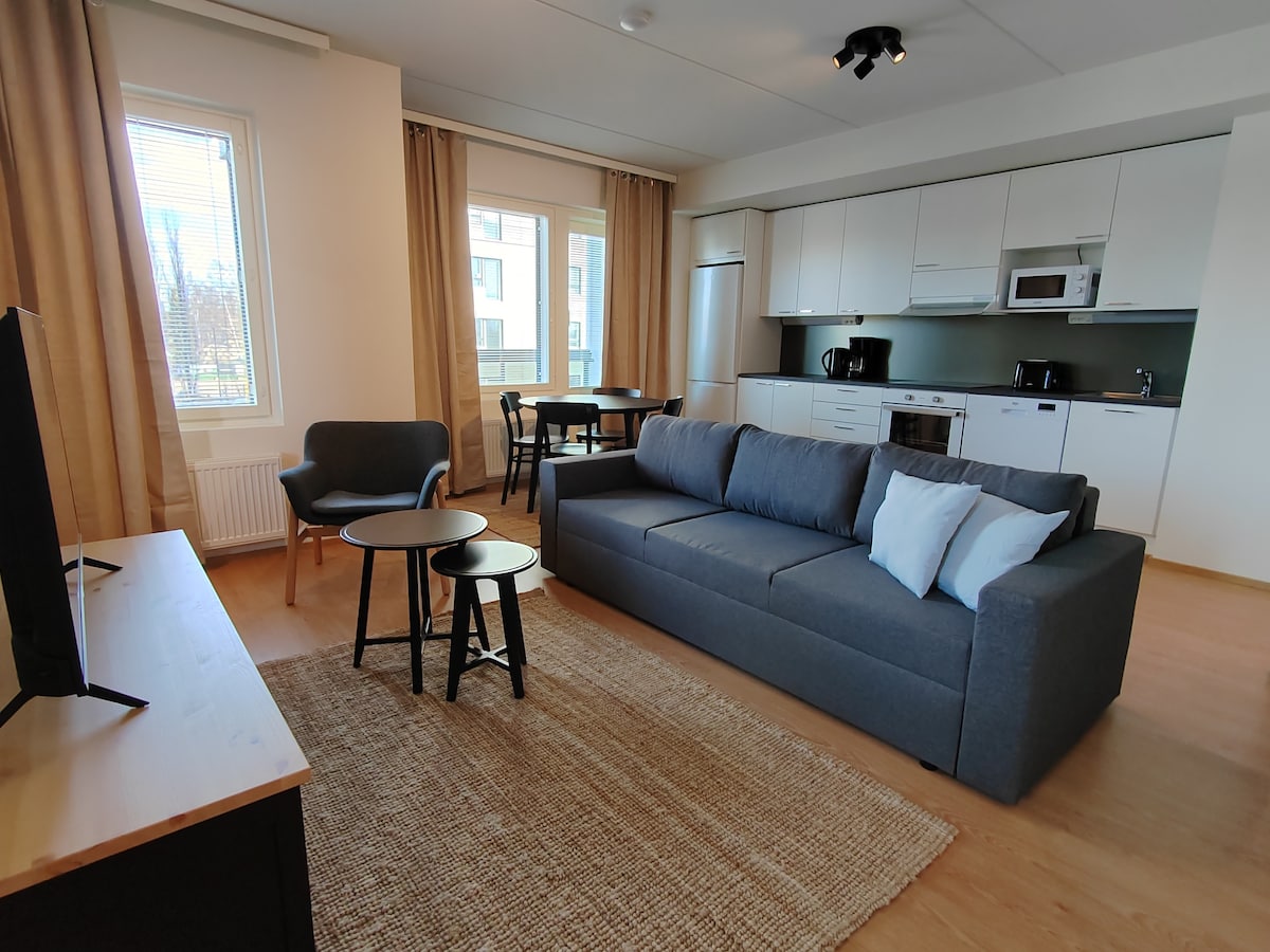 A modern living space is shown, featuring a gray sofa with light accent pillows and a pair of round side tables. Nearby, a dining table accompanies four chairs. The open kitchen includes white cabinetry and essential appliances, with natural light filtering through large windows.