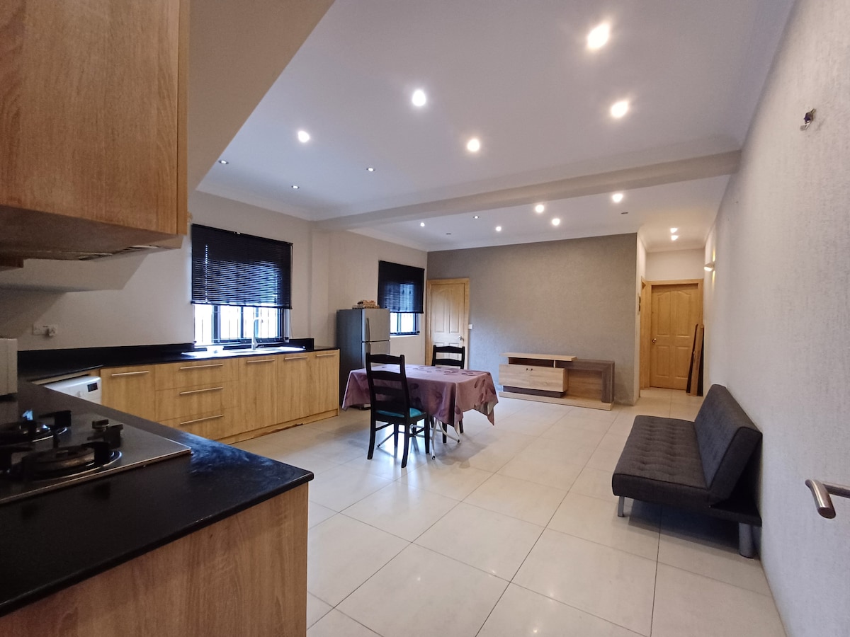A spacious kitchen and living area are depicted, featuring light-colored walls and tiled flooring. A table with chairs occupies the center, while a sleek sofa extends along one wall. Natural light filters through the covered windows, complementing the modern cabinetry and appliances.