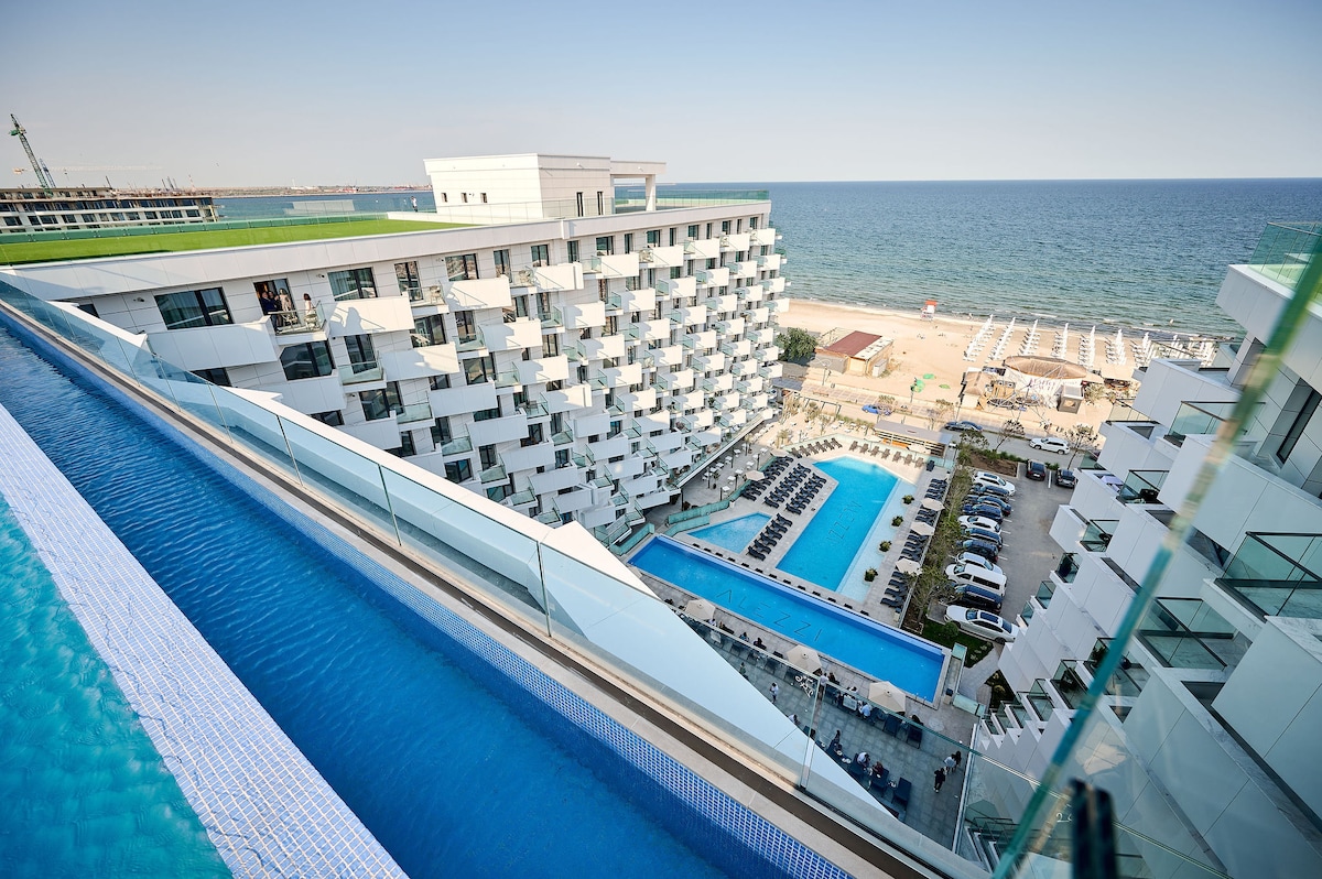 An expansive view from a balcony captures the resort's layout, featuring two swimming pools and the nearby beach. The modern architecture of the building is complemented by lounge chairs arranged along the sandy shore, with the sea stretching beyond the horizon.