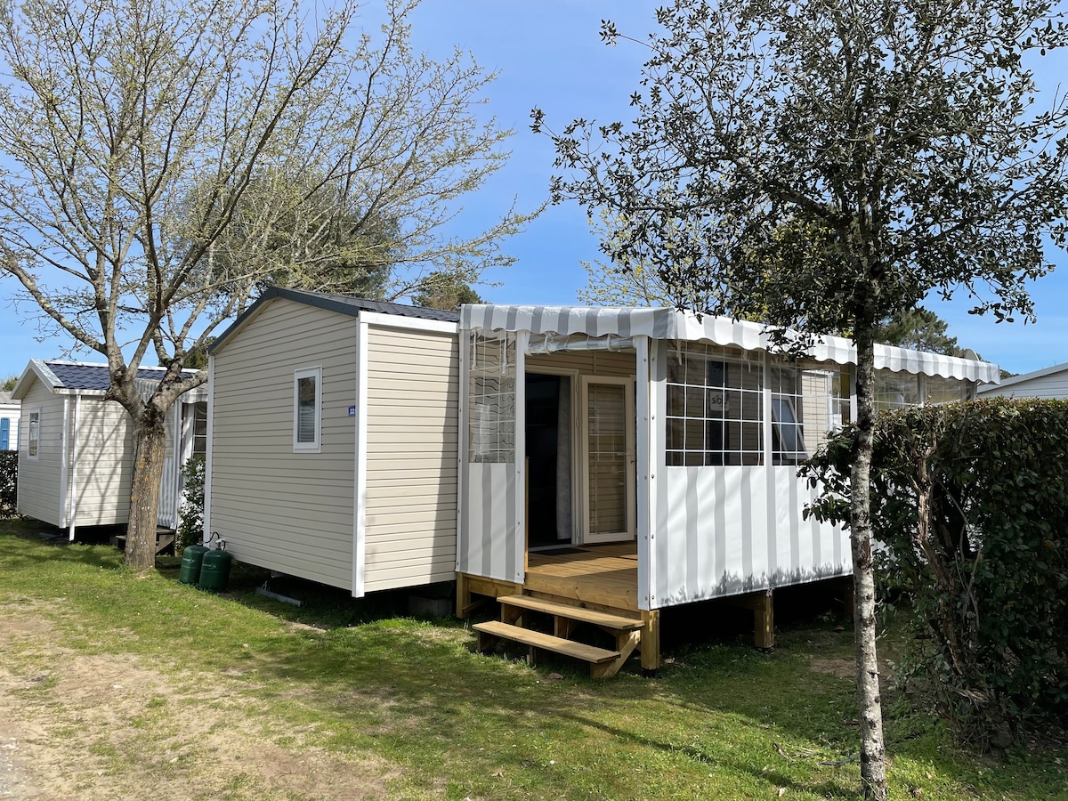 A mobil home is set within a peaceful, tree-lined area. The entrance features a spacious wooden deck, enclosed by a white awning and large glass windows, allowing for natural light to enter. Surrounding greenery enhances the tranquil environment.