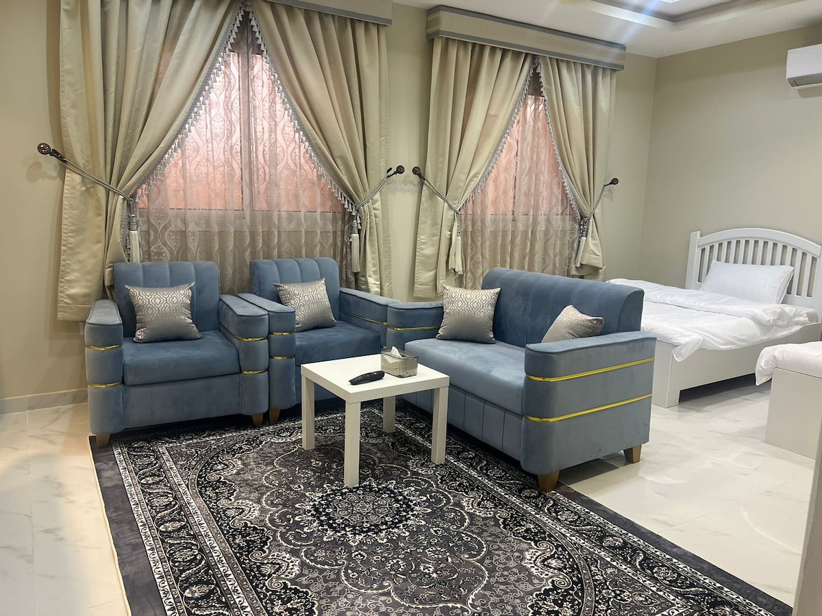 A spacious living area is furnished with two soft blue armchairs and a small white table. Elegant drapes frame large windows, allowing natural light to softly illuminate the room. In the background, a bed is neatly made, contributing to a calm and serene environment.