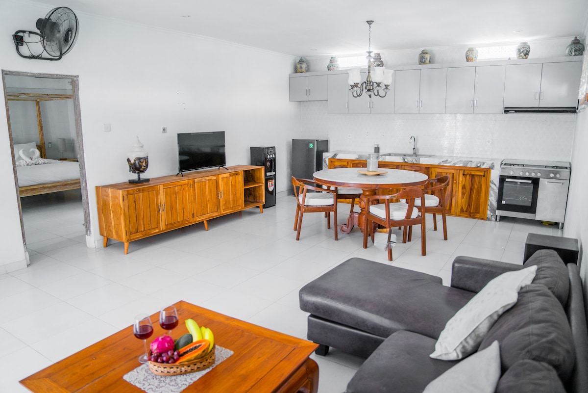 A spacious living area is presented with a comfortable dark sofa and a wooden dining table surrounded by four chairs. A modern kitchen is visible in the background, equipped with appliances and cabinetry. Natural light brightens the white-tiled floor and walls.