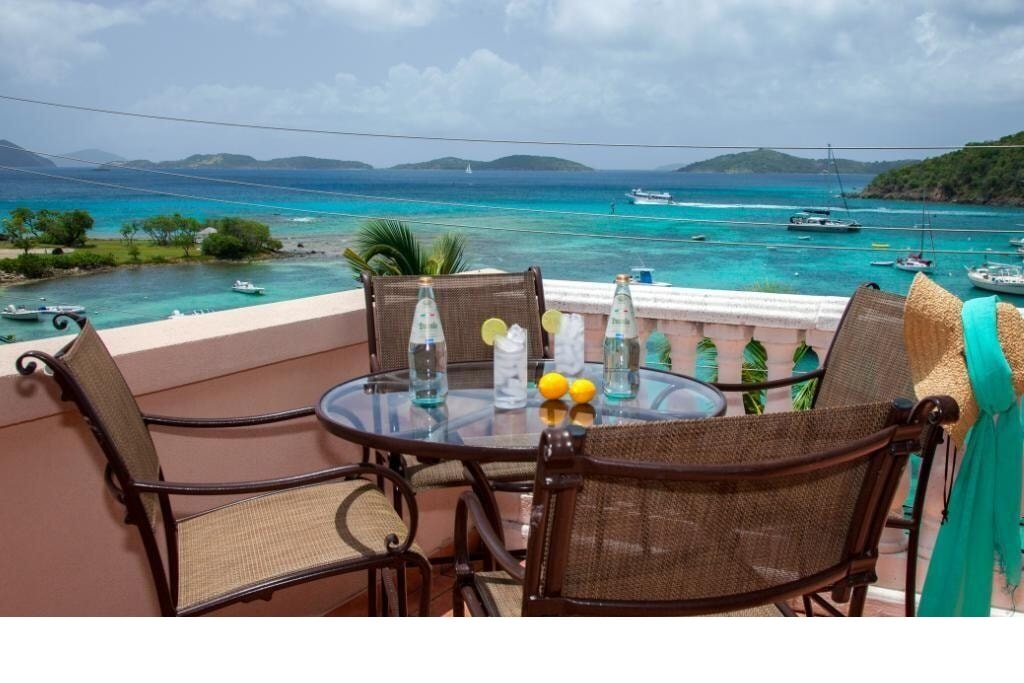 An outdoor dining area features a glass table surrounded by four wicker chairs. Brightly colored lemons and water bottles sit on the table, while the stunning view of Cruz Bay Harbor showcases turquoise waters and anchored boats, with lush islands in the distance.