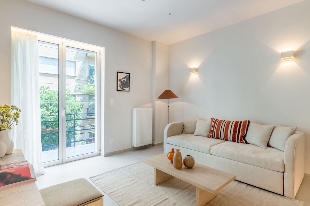 A light-filled living area features a sleek sofa adorned with decorative pillows. A wooden coffee table is positioned in front, with subtle vases and books. Large glass doors open to a balcony, framed by flowing white curtains, and softly lit walls enhance the serene ambiance.
