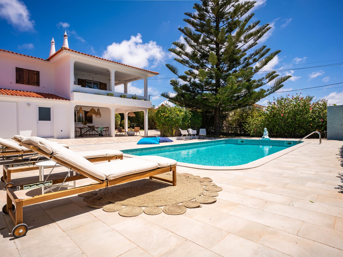 A spacious outdoor area features a swimming pool surrounded by lounge chairs. Lush greenery provides privacy, while a large tree casts shade nearby. The house's balcony is visible above, with inviting outdoor seating beneath it, creating a serene setting for relaxation.