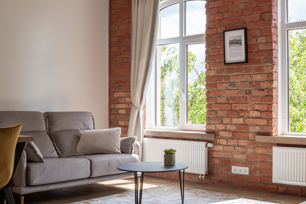A bright living area features exposed brick walls and large windows providing natural light. A comfortable gray sofa is paired with a low coffee table. A small green plant sits on the table, complementing the warm tones of the space.