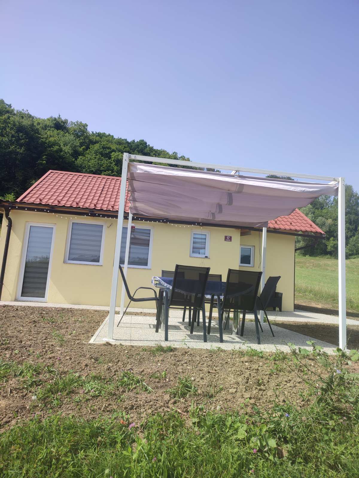 A yellow house features a red tiled roof against a backdrop of lush greenery. An outdoor dining area with a shaded pergola provides a setting for meals, equipped with six dark chairs around a rectangular table. The surroundings display open natural space.