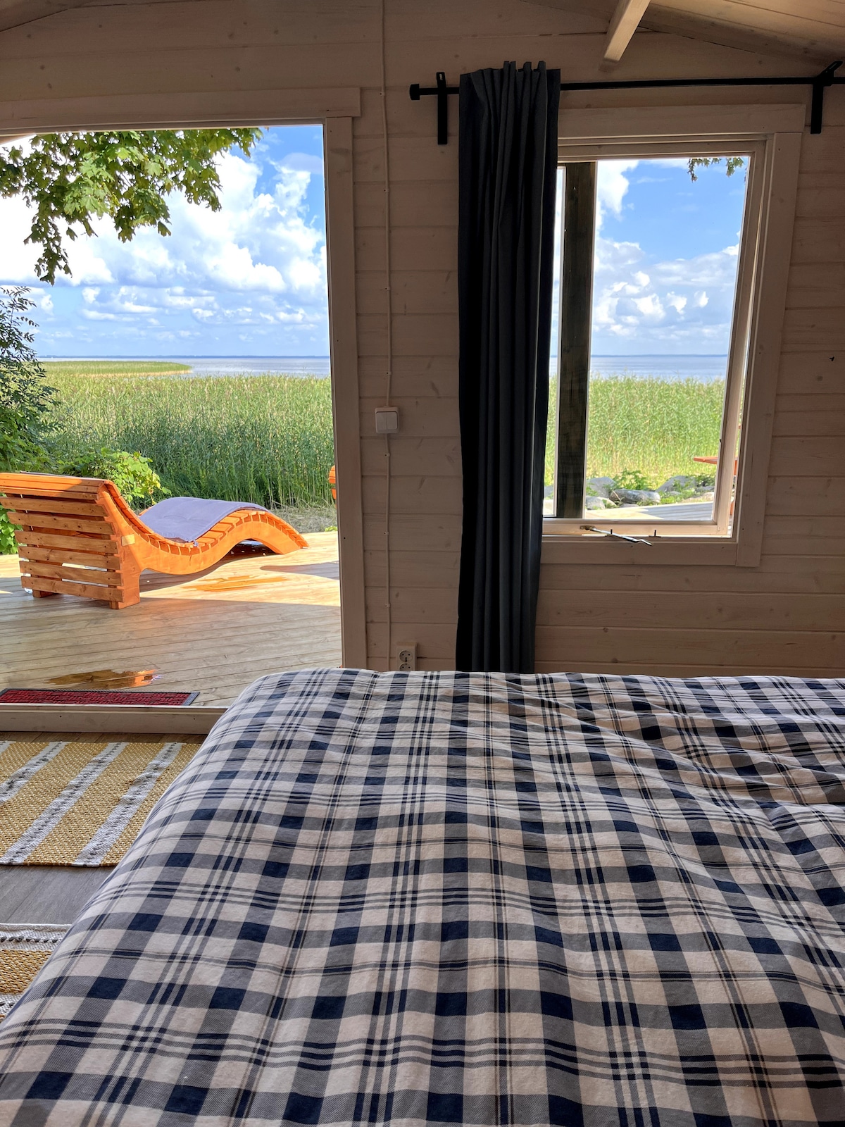 A cozy sleeping area is shown with a checked blanket draped over a spacious bed. A window reveals a scenic view of lush greenery and water beyond. Natural light fills the room, with a comfortable lounge chair visible on the terrace outside.