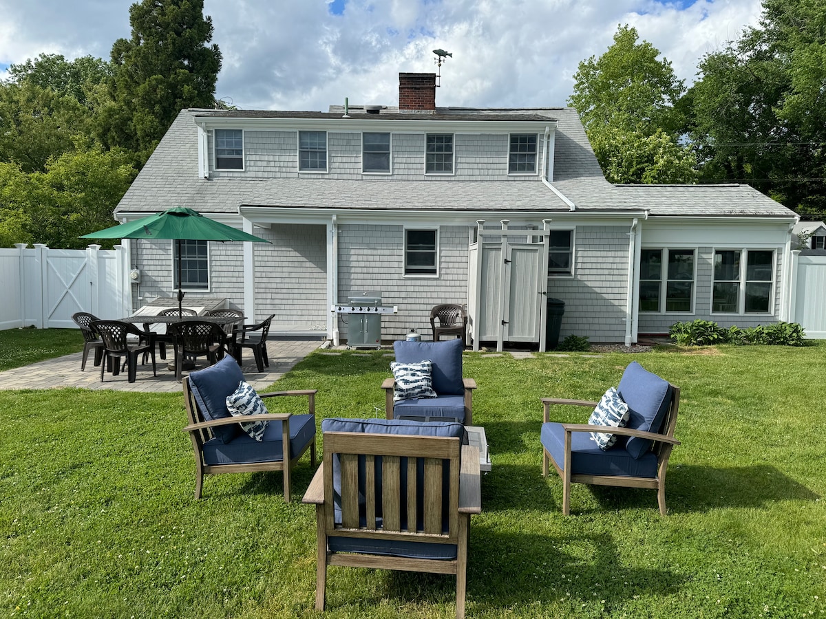 The outdoor area features a well-maintained lawn with a seating arrangement of four chairs and a table surrounded by greenery. A large umbrella provides shade over the space. The house displays a neutral exterior with multiple windows and access to a backyard privacy fence.