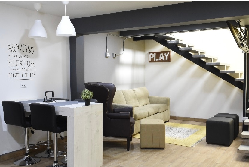 A modern living area features a cozy sofa in a neutral tone, complemented by a patterned rug. A dining table with high stools occupies one corner, while the staircase, adorned with a wooden sign reading 'PLAY', adds character to the space.