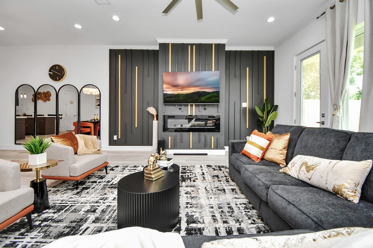 A contemporary living room is presented, featuring a large sectional sofa adorned with decorative pillows. A sleek black coffee table sits atop a patterned area rug. A mounted television is surrounded by a striking wall design, while large windows provide natural light and a view of the outdoors.