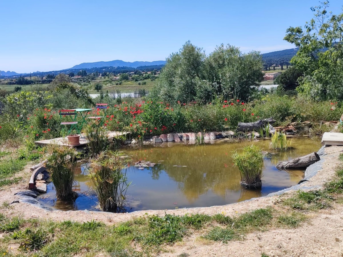A tranquil pond is framed by lush greenery and flowering plants, reflecting the surrounding landscape. Natural materials, including stones and logs, enhance the organic feel of the space. Scenic hills and distant mountains are visible in the background, creating a peaceful atmosphere.