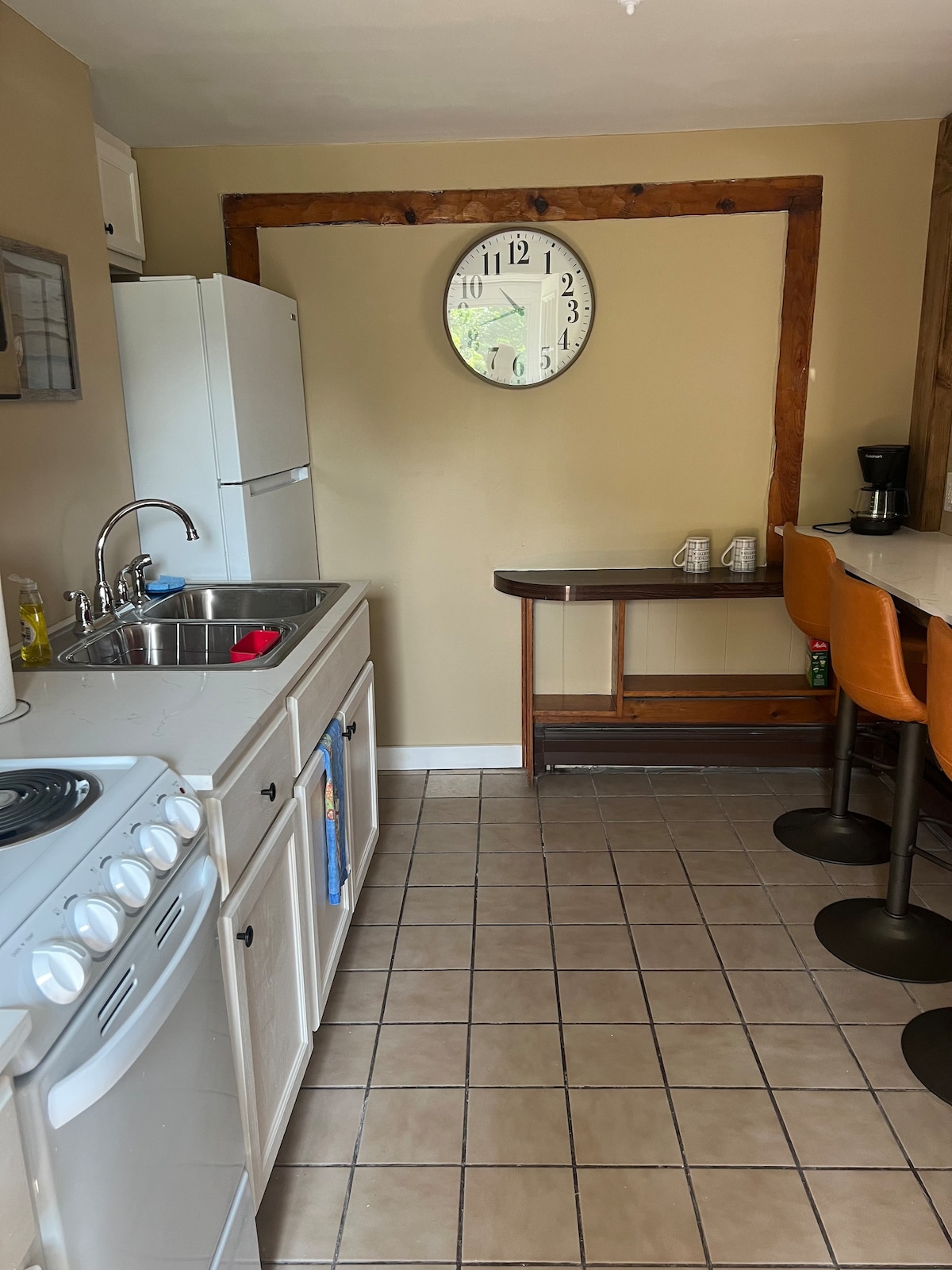 A compact kitchen area is equipped with essential appliances, including a stove and refrigerator. A bar counter with two orange stools offers additional seating. A large wall clock is mounted above the sink, and a window brings in natural light. Neutral-toned walls complement the tiled floor.