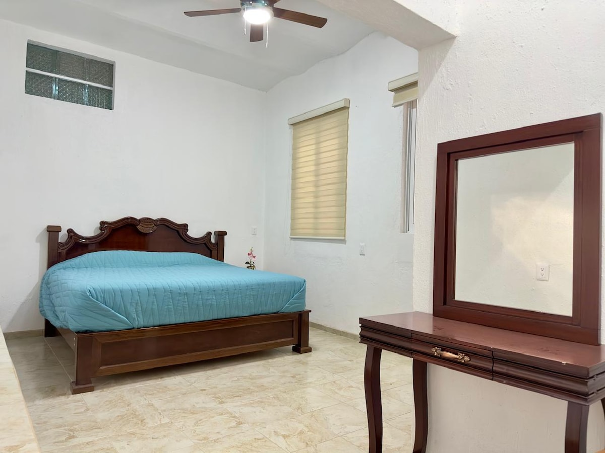 A spacious sleeping area features a wooden bed with a blue coverlet. Natural light enters through nearby windows adorned with light shades. A wooden vanity table is positioned against one wall, complemented by a ceiling fan for airflow.