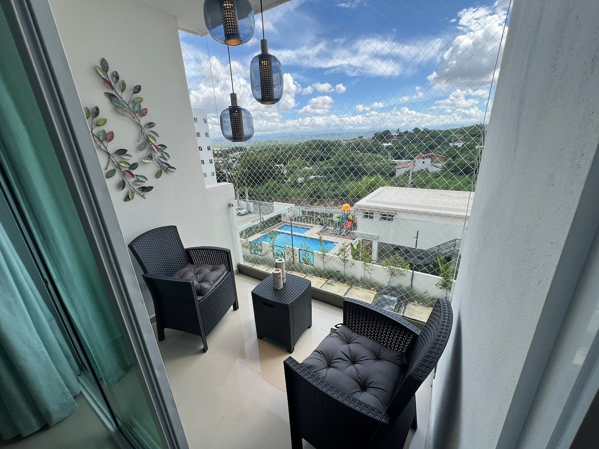 A balcony area is equipped with two black wicker chairs and a small table, offering a view of the outdoor pool below. Modern pendant lights hang overhead, and a decorative wall piece featuring leaves enhances the space. Lush greenery and the sky can be seen in the background.