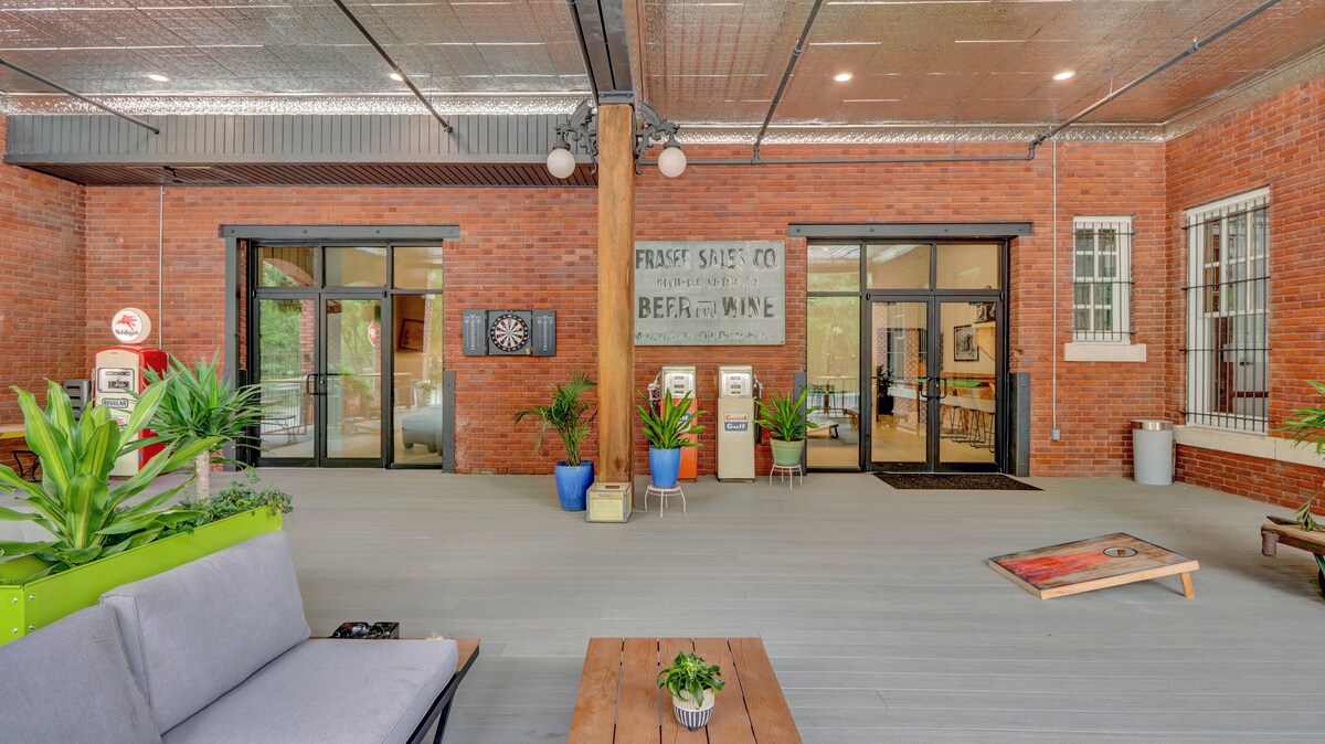 An open and inviting common area is showcased with a textured ceiling and large glass doors. Planters with greenery are placed throughout, and a dartboard is mounted on the wall. Seating is arranged on a wooden deck, providing a relaxed atmosphere for socializing.
