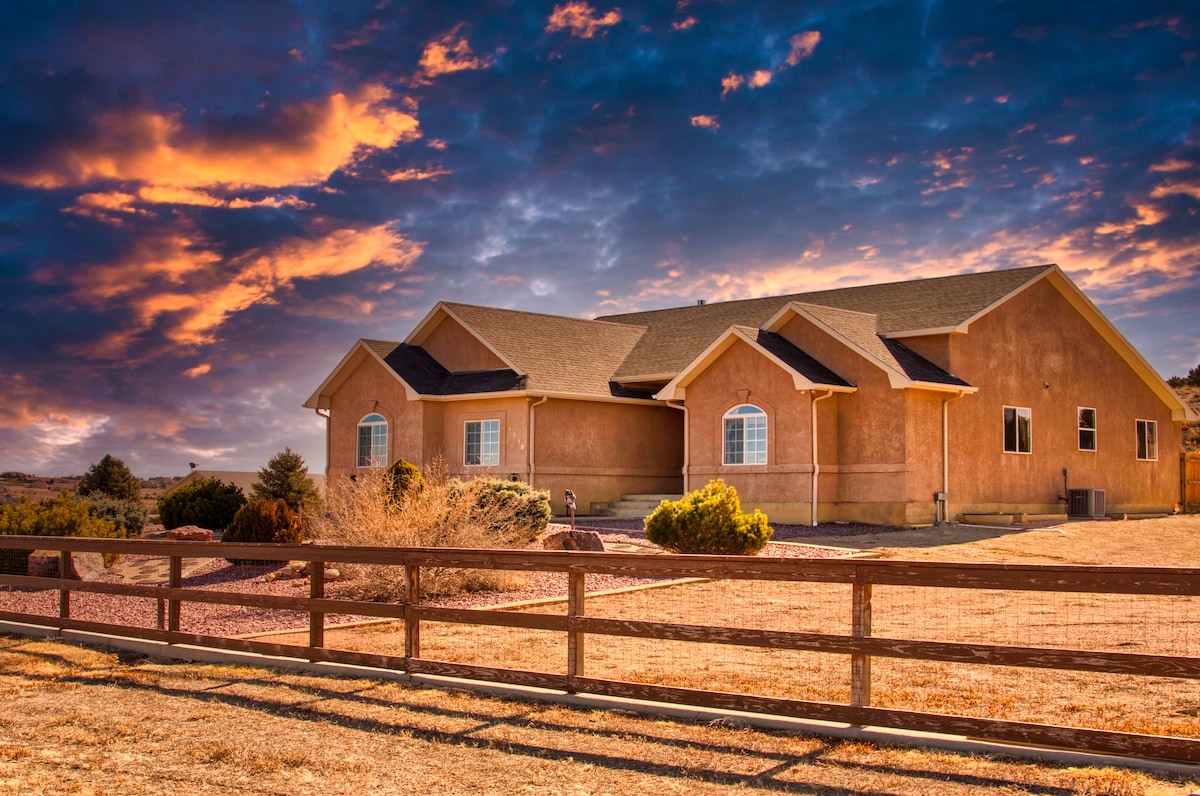 A spacious house is depicted against a dramatic sunset sky, showcasing a warm exterior and multiple windows. The surrounding landscape features dry grass and scattered shrubs, with a wooden fence framing the front yard.