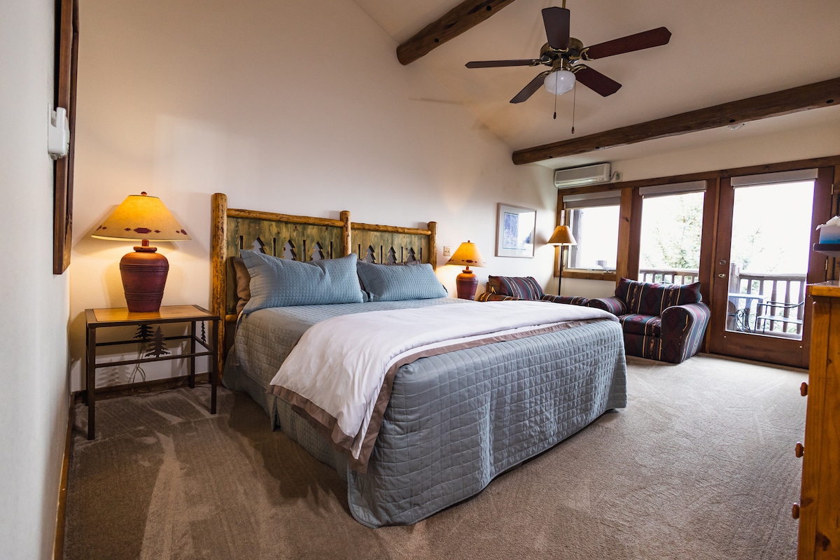 A spacious guest room features a king bed with a textured blue coverlet, complemented by rustic wooden bed posts. A ceiling fan hangs above, and two bedside lamps illuminate the room. Comfortable seating is offered with a cozy loveseat and chair, and large doors provide access to a private deck.