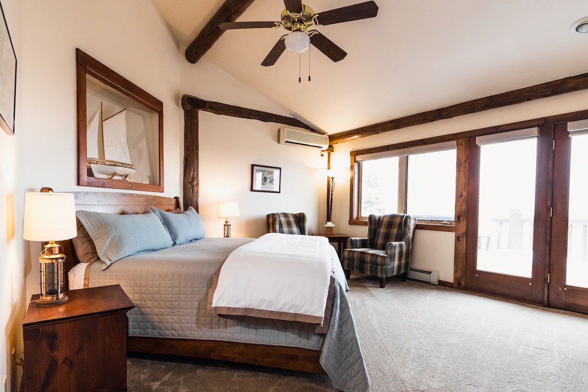 A spacious bedroom features a king bed covered with a light quilt, accompanied by a side table with a lamp. A cozy armchair is positioned near large windows, providing natural light. Exposed wooden beams and a ceiling fan add rustic charm to the space.