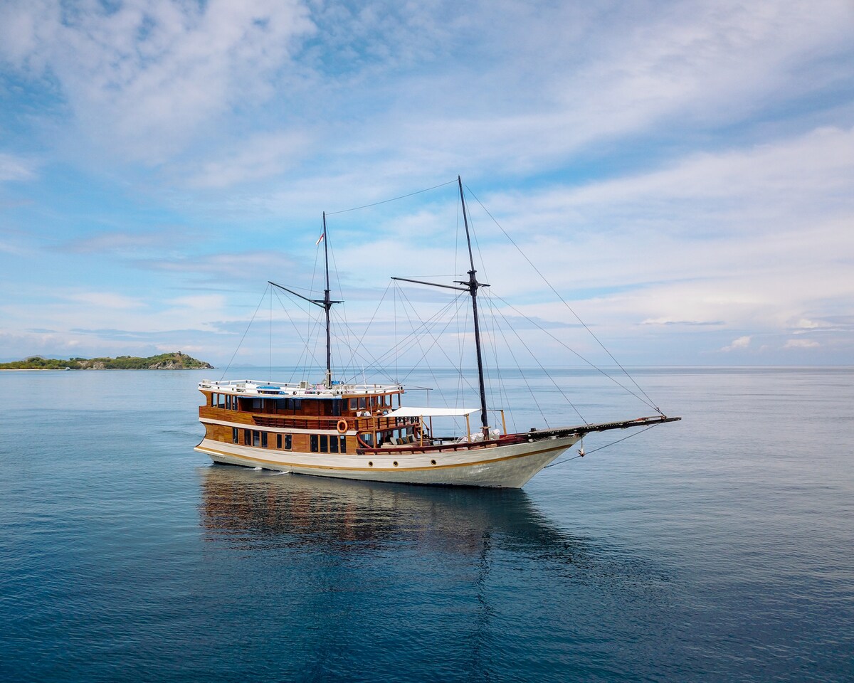 A deluxe phinisi boat is anchored on calm waters, showcasing its wooden structure and two tall masts. The serene backdrop highlights a clear sky with scattered clouds, emphasizing the tranquility of the surroundings.