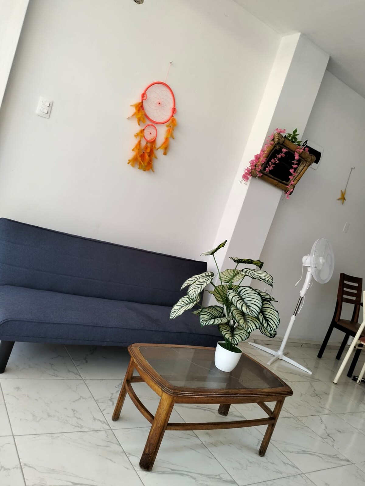 A compact living area features a dark blue sofa and a glass-topped coffee table, complemented by a vibrant plant in a white pot. A wall hanging of dreamcatchers adds a decorative touch, while a fan and wooden chairs provide functional elements to the space.