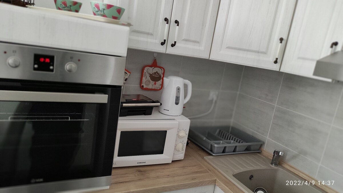 A modern kitchen features a combination of stainless steel and white cabinetry. An oven and microwave are prominently displayed. A kettle sits on the counter, alongside a dish rack and a sink with a minimalist design. Decorative bowls are placed on the upper shelf.
