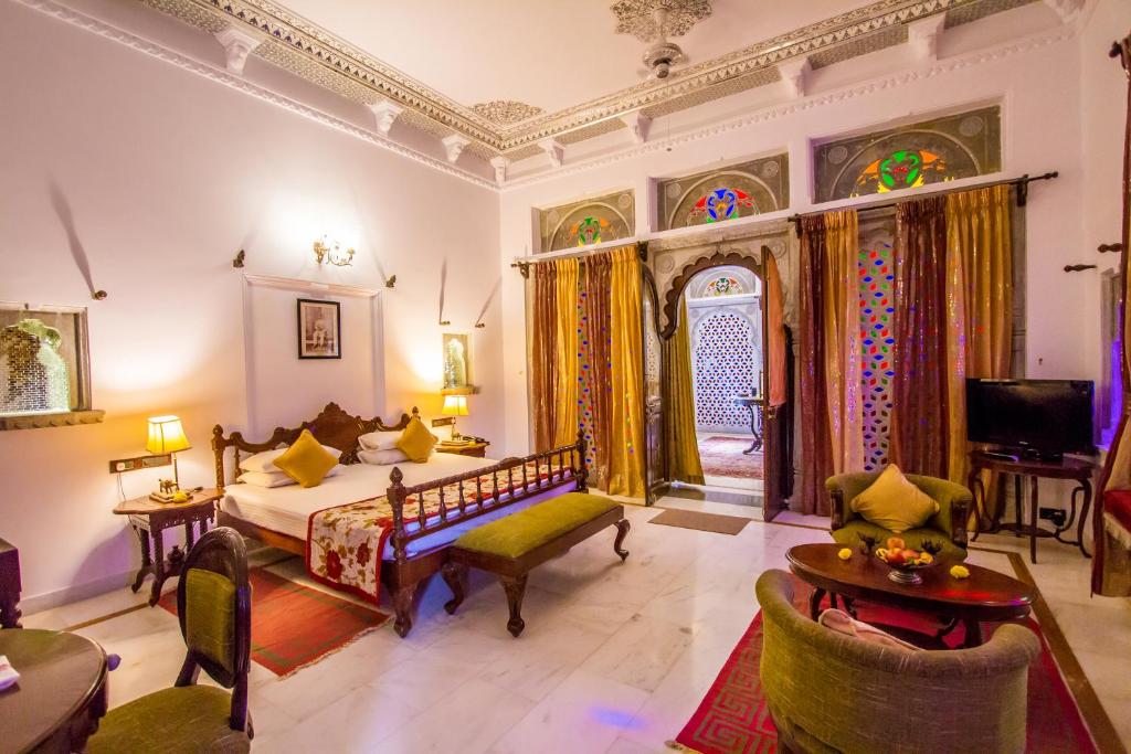 A spacious room features a warm color palette, with ornate decor and traditional furnishings. A large bed is positioned prominently, complemented by a seating area with green chairs and a wooden table. Colorful stained glass windows add texture and interest, enhancing the natural light.
