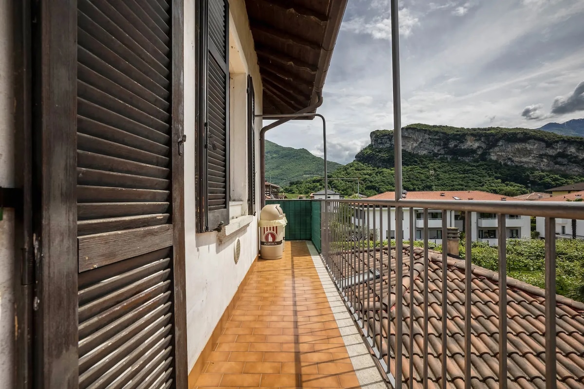 A balcony with a tiled floor extends outward, offering unobstructed views of the surrounding mountains. Wooden shutters frame the entrance, while a small storage area is visible at one end. The landscape features lush greenery and rooftops, creating a serene outdoor space.
