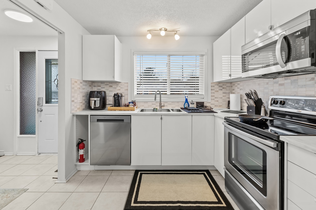 The kitchen features modern white cabinetry and stainless steel appliances. A sink is centrally located under a window, allowing natural light in. A stove, microwave, and coffee maker can be seen, along with a wooden cutting board. Tile flooring is complemented by a geometric area rug.