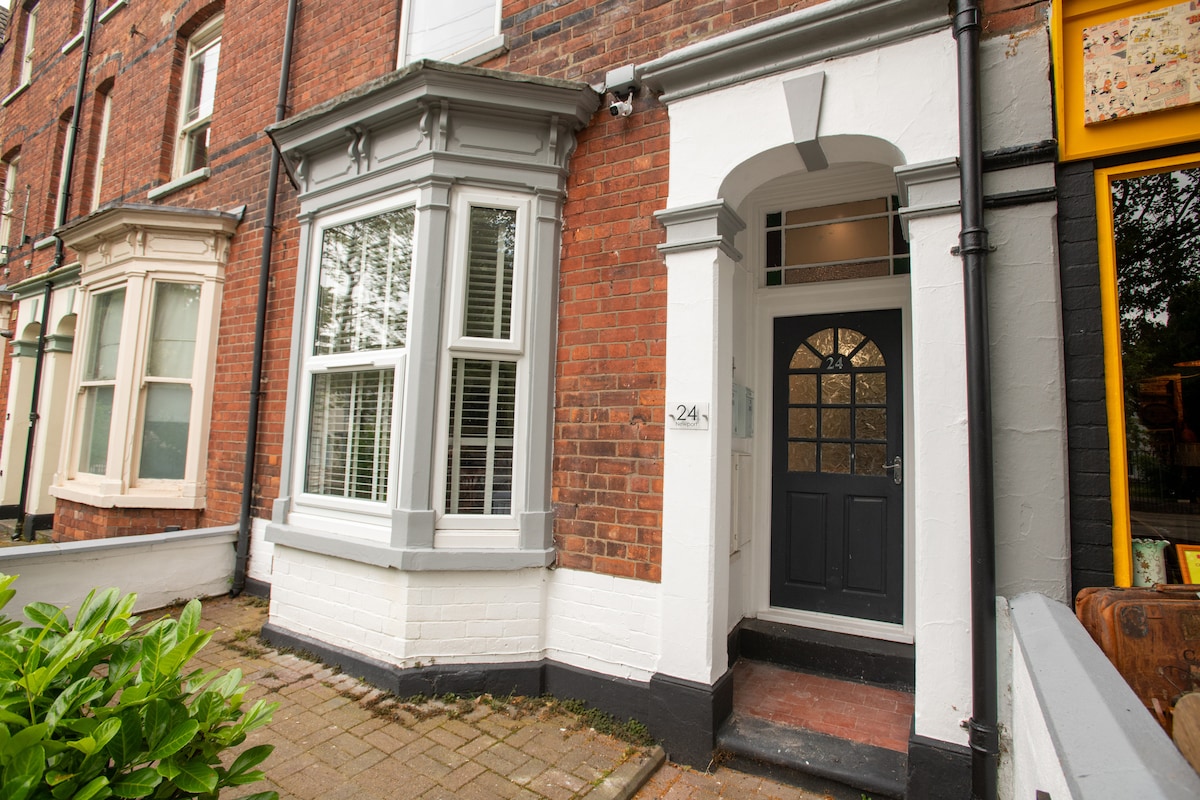 The exterior of the apartment is shown, featuring a brick facade with white trim. A welcoming entrance is highlighted by a dark door, complemented by large windows that allow natural light. The surrounding greenery and pathway contribute to a pleasant approach.