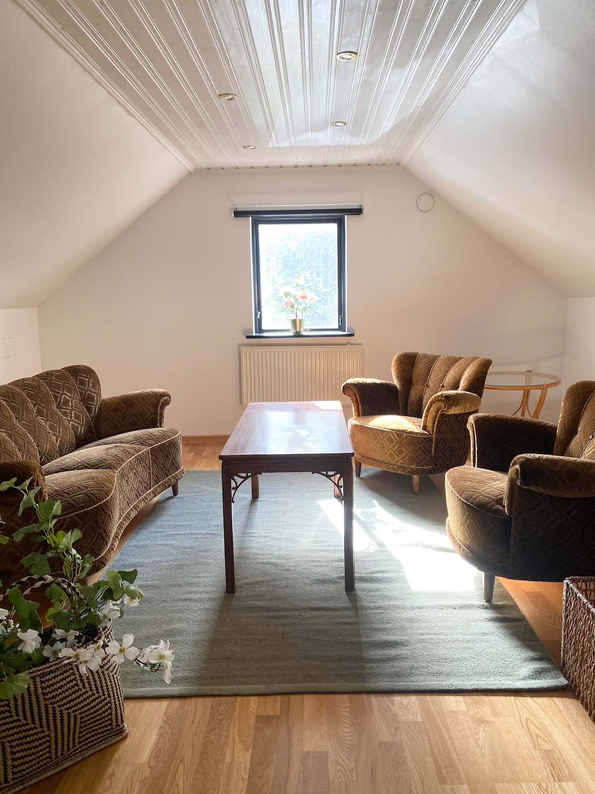 A comfortable seating area features two armchairs and a sofa, arranged around a wooden coffee table. Natural light filters through a window adorned with light-filtering shades, highlighting the textured surfaces. A small side table rests beside an armchair, with decorative plants in a woven basket adding a touch of greenery.