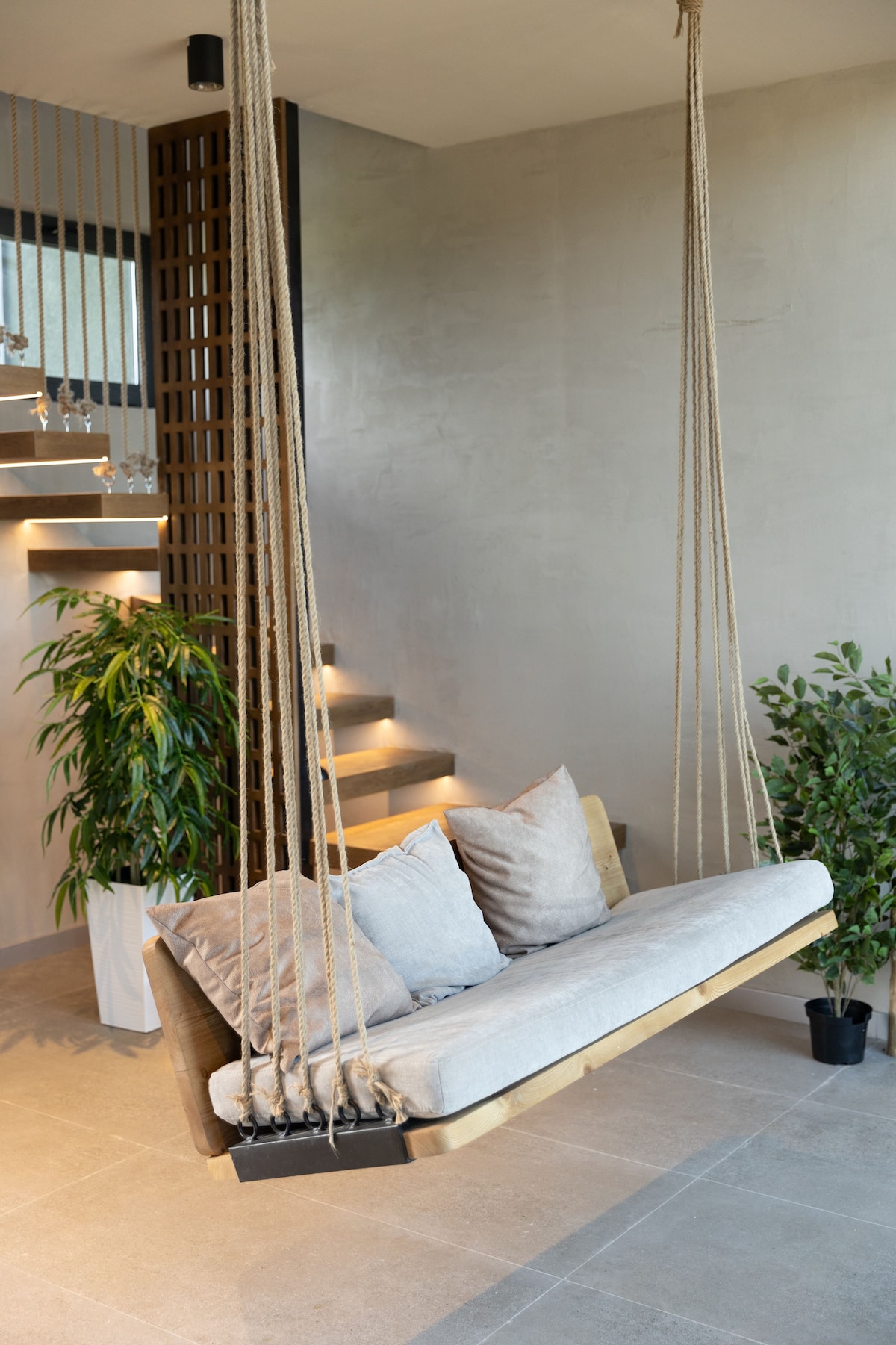 A suspended wooden swing seat is positioned in a modern living area, adorned with soft cushions. Lush green plants create a refreshing contrast, while warm lighting highlights the smooth concrete walls and stylish staircase in the background.