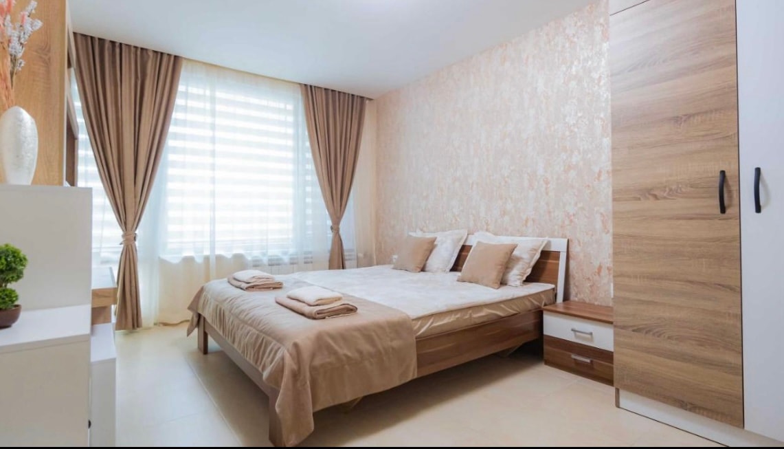 A spacious bedroom features a large bed dressed in neutral linens, flanked by soft decorative pillows. Natural light filters through sheer curtains over a window. A wooden wardrobe and a smaller nightstand are present, enhancing the overall warmth of the space.