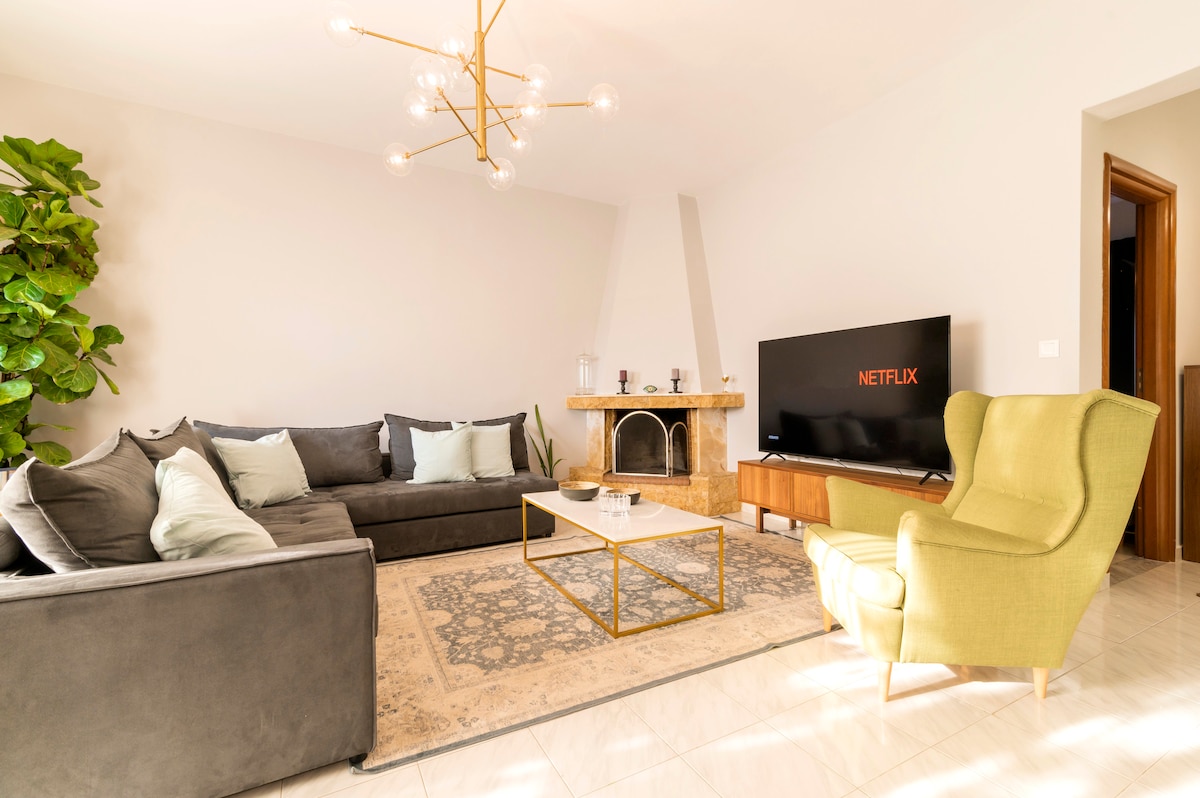 A living room is presented with a sectional sofa and an accent chair. A coffee table sits on a soft area rug, accompanied by a decorative plant. A TV mounted on the wall displays Netflix. Natural light filters through the room, enhancing its open layout.