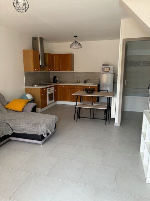 A modern kitchen area features wooden cabinetry and stainless steel appliances. A dining table with two chairs is positioned beside a cozy, spacious couch adorned with a grey throw and a colorful pillow. Neutral-toned tiles cover the floor, providing a clean and open look.