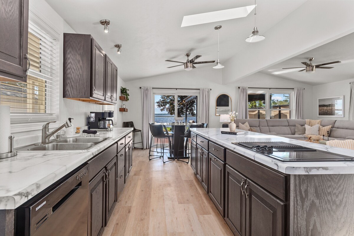 A modern kitchen is shown with dark wood cabinetry and marble countertops, complemented by stainless steel appliances. Natural light filters through skylights above, illuminating the open space that connects to a casual dining area featuring seating and views of the lake.
