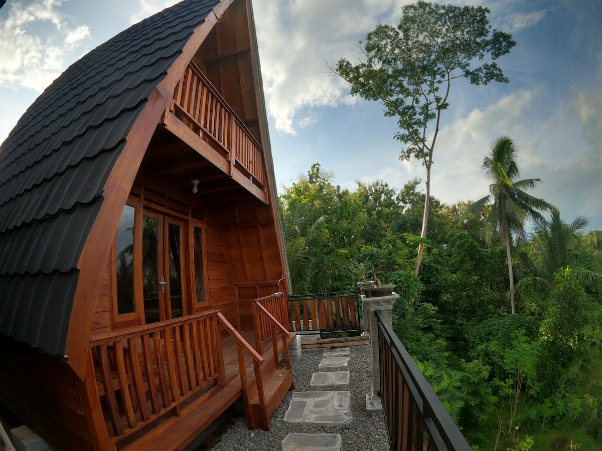 The cabin features a striking angled roof and wooden exterior, surrounded by lush greenery. A stone path leads to the entrance, which is framed by large windows allowing natural light to fill the space, and a balcony offers views of the tropical landscape.