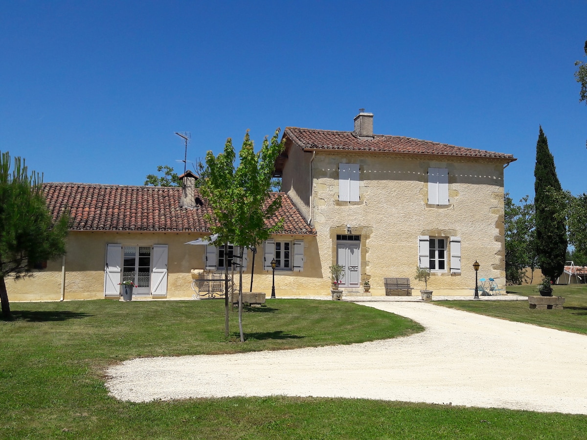 A charming house with a stone facade and a tiled roof is surrounded by a grassy area. White window shutters complement the exterior. A gravel driveway leads to the entrance, with a small seating area visible on the patio.