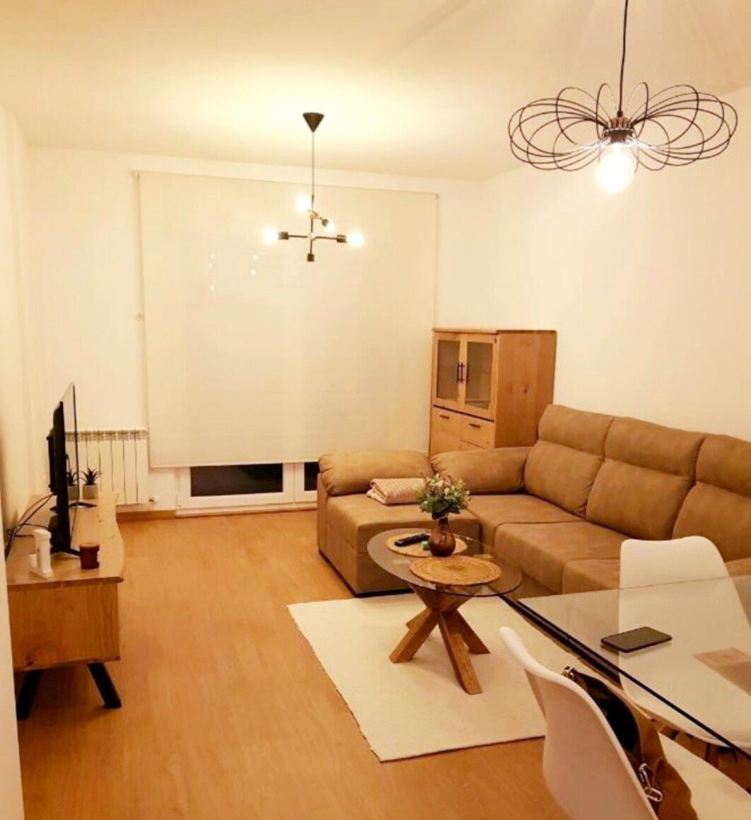 A living area features a sectional sofa in a neutral tone, complemented by a wooden coffee table. A TV is positioned against the wall. Natural light enters through a large window dressed with a light blind, enhancing the bright and airy feel of the space.